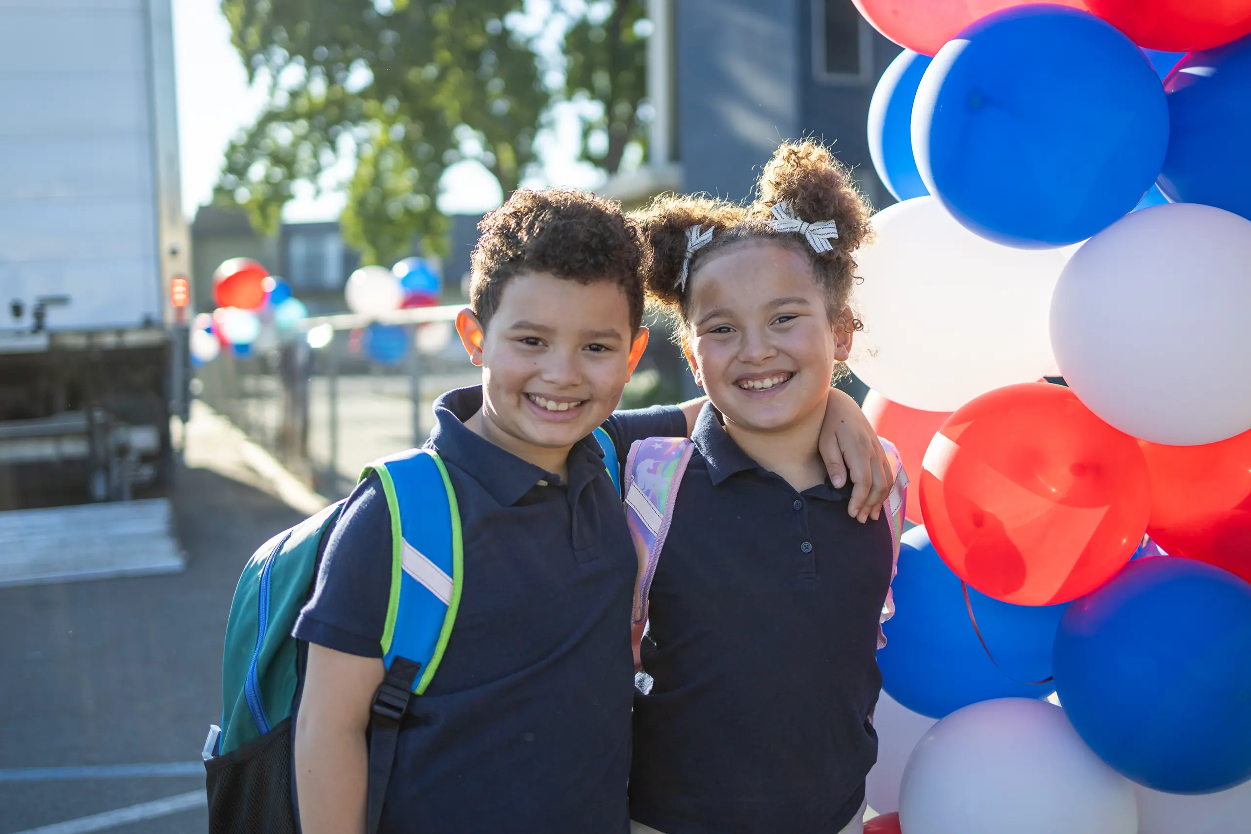 Two smiling children with backpacks stand arm-in-arm next to a cluster of red, white, and blue balloons outdoors, likely at a school event.